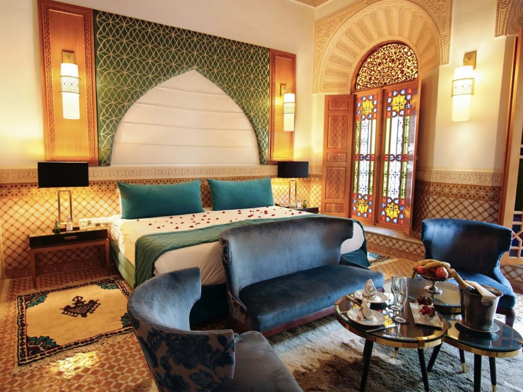 A suite bedroom at Riad El Amine Fès with a green zellige-patterned headboard, teal velvet pillows and rose petals scattered across the bed, a stained-glass window glowing beside blue velvet armchairs and a champagne bucket set for arrival.