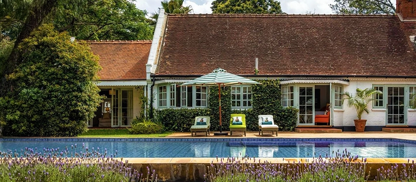 The white-rendered facade of Hamilton House viewed across the swimming pool, its terracotta-tiled roof and sage-green French doors framed by lavender borders and lush tropical gardens