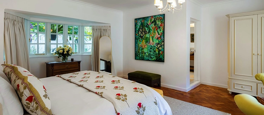 A light-filled guest bedroom at Hamilton House featuring a king bed with a floral bedspread, parquet floors, a large vivid green botanical artwork and a chandelier above