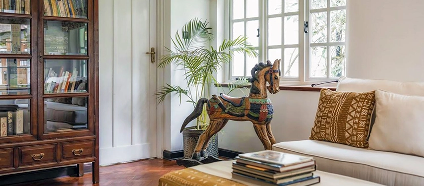 A living room corner at Hamilton House with an antique glazed bookcase, a painted carved wooden rocking horse, a cream sofa with an African mudcloth cushion and white-framed windows