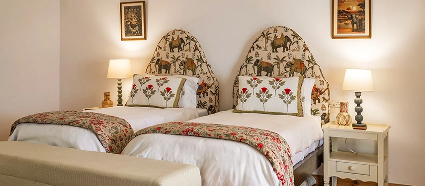 The twin bedroom at Hamilton House with two beds featuring arched headboards upholstered in elephant-print fabric, matching floral bedspreads and framed vintage African artwork on the walls