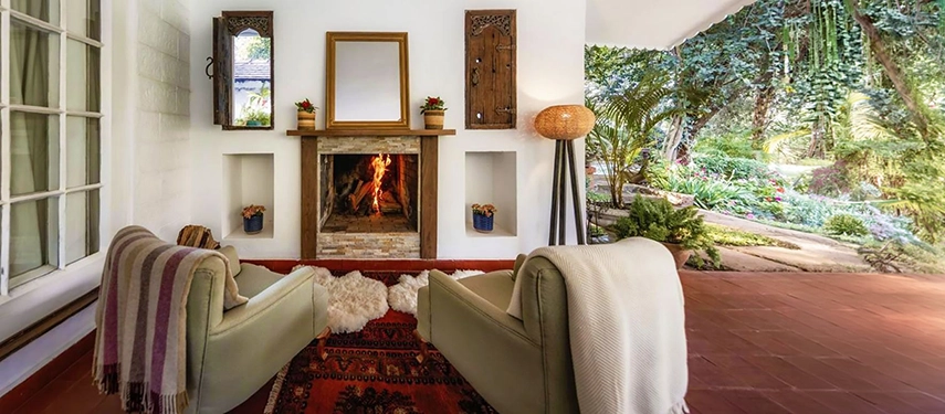 A veranda sitting area at Hamilton House centred on a lit stone fireplace, with two green armchairs draped in throws, a kilim rug underfoot and garden greenery visible beyond