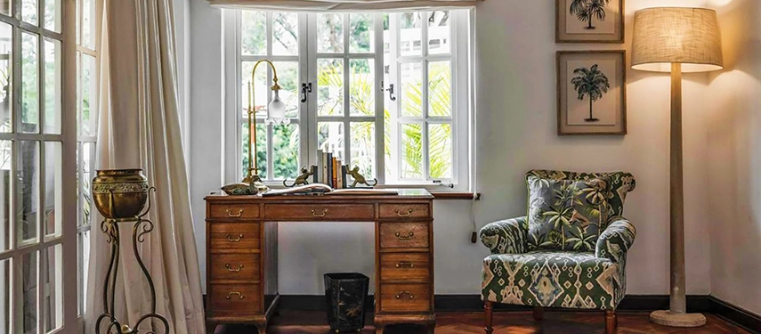A study corner at Hamilton House featuring an antique mahogany pedestal desk with a brass lamp, a botanical-print armchair and framed palm illustrations above a white-framed bay window
