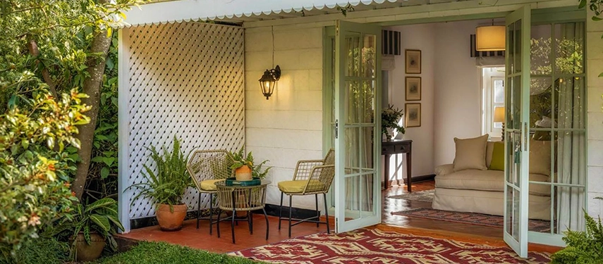The private veranda of the Acacia Room at Hamilton House, with rattan chairs and a small table set against a whitewashed lattice wall, sage-green French doors open to the room beyond