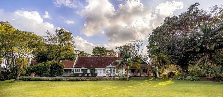 Hamilton House viewed across its wide, manicured lawn under a dramatic sky, the whitewashed 1930s colonial house flanked by towering trees and lush tropical gardens