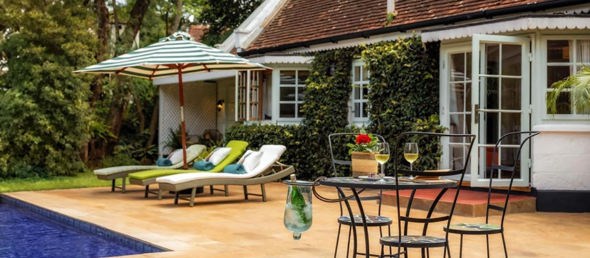 The pool terrace at Hamilton House with sun loungers, a striped umbrella and a bistro table set with wine glasses, the ivy-clad white facade and sage-green French doors behind
