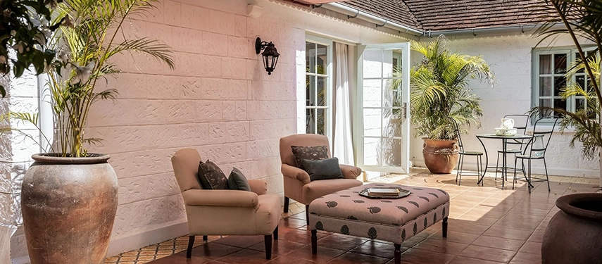 A sunlit courtyard seating area at Hamilton House with two armchairs and an upholstered ottoman, large terracotta urns planted with palms and French doors opening to the rooms beyond
