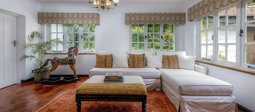 The Drawing Room at Hamilton House with a deep white corner sofa, an embroidered ottoman, African mudcloth cushions, parquet floors and a carved wooden rocking horse beside tall windows