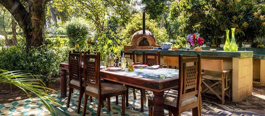 The outdoor dining and pizza oven area at Hamilton House, a long wooden table set with bottles and flowers beneath canopied trees, a domed wood-fired oven visible in the background