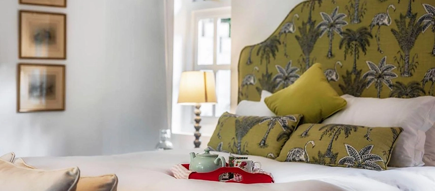 A close view of a bed at Hamilton House with an olive-green palm-print headboard, matching cushions and a red lacquer breakfast tray with a celadon teapot
