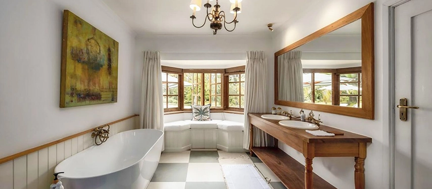 A generous ensuite bathroom at Hamilton House with a freestanding bath, a wooden double-vanity beneath a large framed mirror, a chandelier and a bay window overlooking the garden