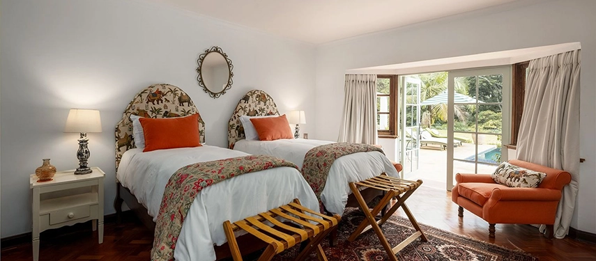 A twin bedroom at Hamilton House with two arched elephant-print headboards, orange accent cushions, parquet floors and sliding glass doors opening directly onto the swimming pool terrace