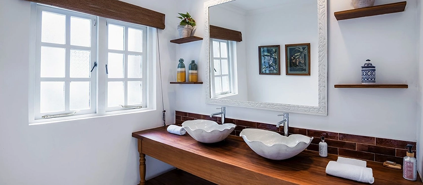 An ensuite bathroom at Hamilton House with two sculpted shell-shaped vessel basins on a wooden vanity, a white ornate mirror, open shelving and small-paned windows