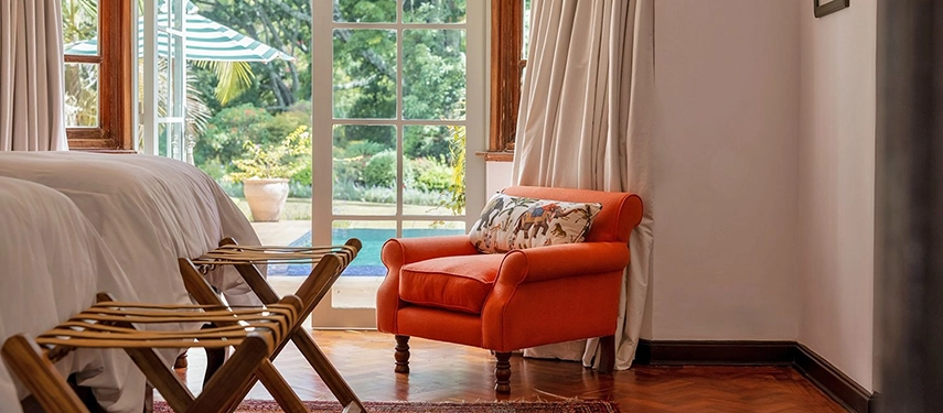 A bedroom detail at Hamilton House showing an orange armchair with a wildlife-print cushion beside tall French windows overlooking the swimming pool and lush garden beyond
