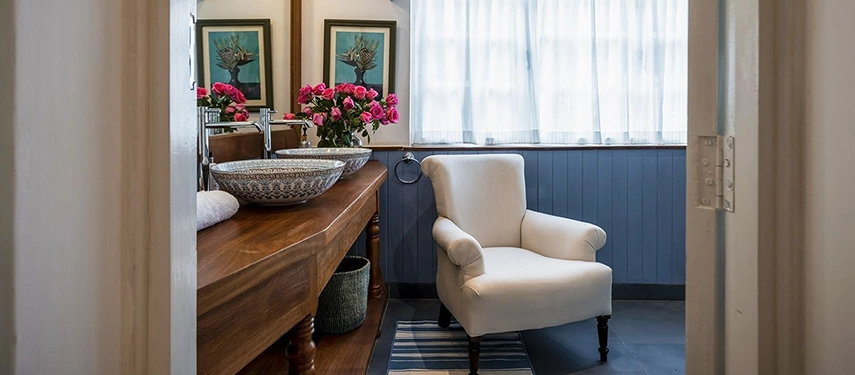 An ensuite bathroom at Hamilton House with a wooden vanity, a patterned ceramic vessel basin, a white armchair, a vase of pink roses and blue-painted timber wainscoting