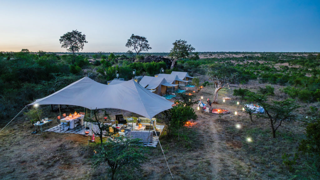 Lantern-lit mobile tented camp at dusk in Laikipia, with a stretch-tent lounge, dining table and guests around a campfire.