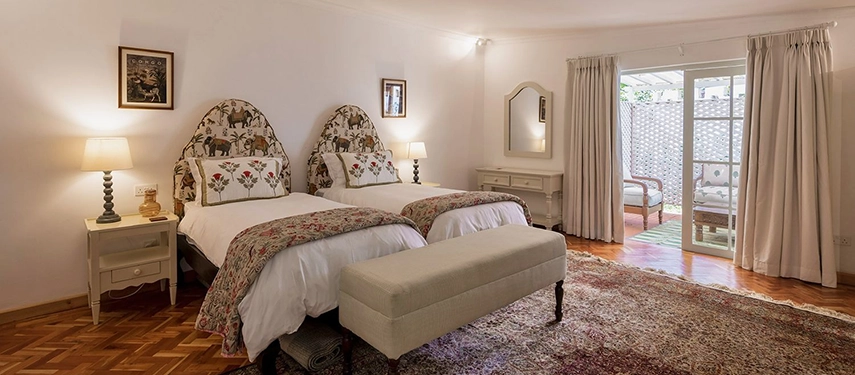 A twin bedroom at Hamilton House with arched elephant-print headboards, floral bedspreads, an upholstered bench, parquet floors and French doors opening to a private veranda beyond