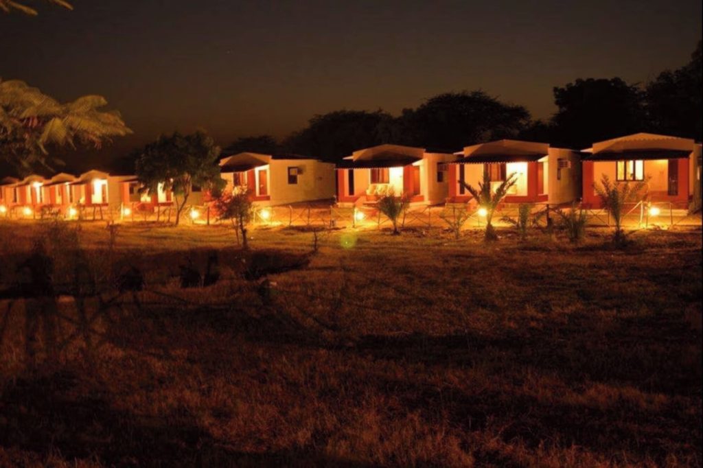 Row of lit cottages with red trim glowing at dusk, Kutch Safari Resort