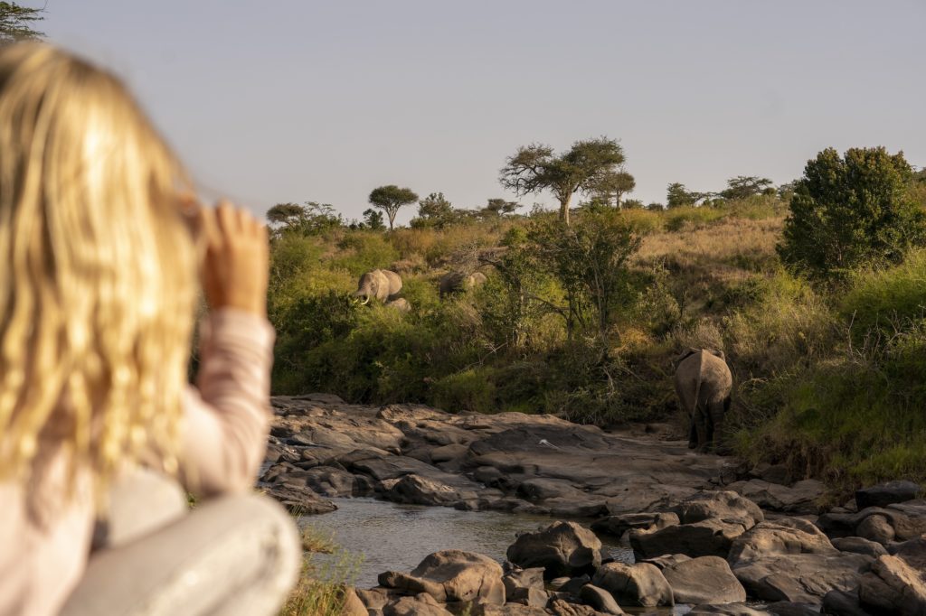 Young girl watching a herd of elephants cross a rocky riverbed in golden evening light at El Karama, Laikipia.