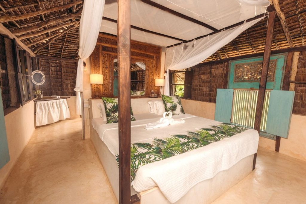Four-poster bed with mosquito net, carved wooden headboard and makuti thatched roof in a bedroom at The Cabanas Lamu.