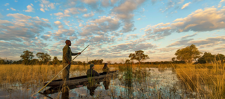 Mokoro safari experience in the Okavango Delta, with guide navigating traditional canoe through tranquil waters, surrounded by lush vegetation and dramatic sunset sky.
