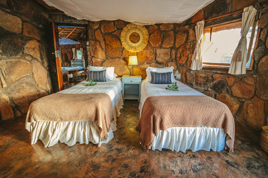 Twin beds with woven throws in a stone-walled bedroom, with a mosquito net overhead and a raffia mirror above the bedside lamp.