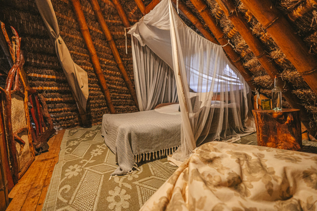 Loft bedroom under a thatched roof, with a netted double bed, a woven floor mat and a tree-trunk bedside table.
