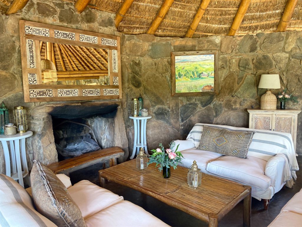 Stone-walled lounge at El Karama Lodge, with an open fireplace, white sofas, a bamboo coffee table and a painted Laikipia landscape.