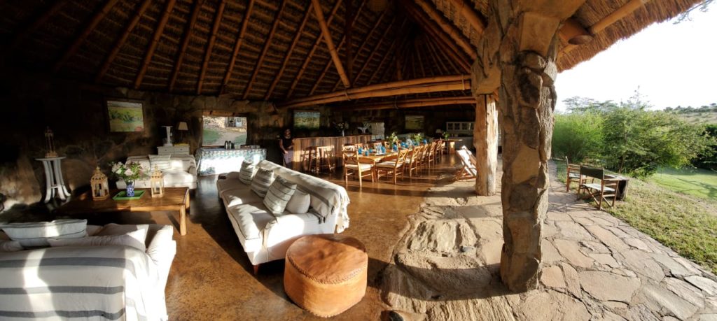 Open-sided thatched lounge and dining area at El Karama Lodge, with white sofas, stone pillars and a long dining table.