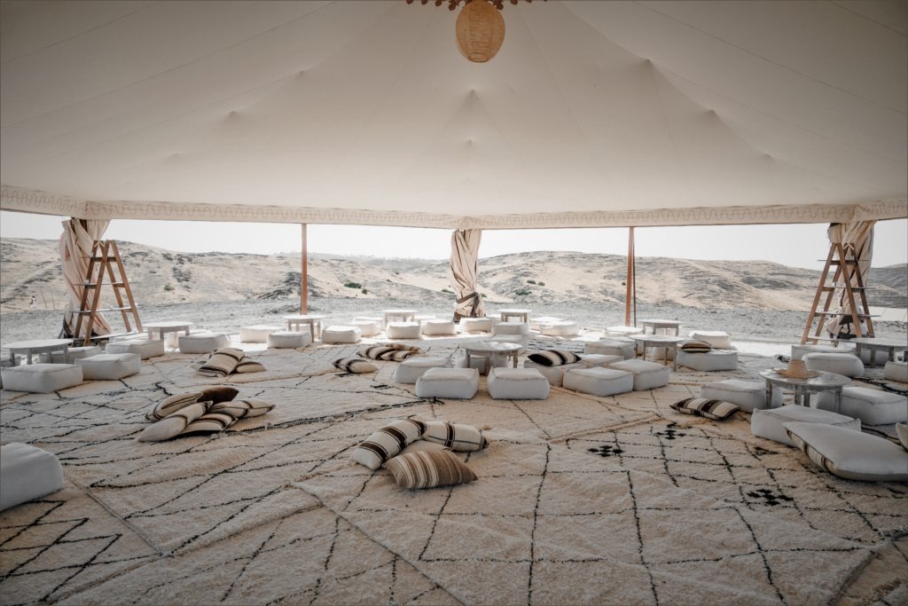 A white canvas tent open to the Agafay hills, its interior scattered with floor cushions and low tables set out for lounging.