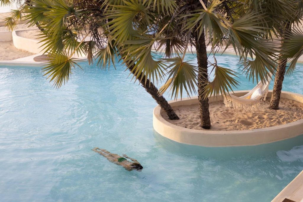 Guest swimming underwater in a turquoise pool around a sandy island planted with palms and a hanging hammock.