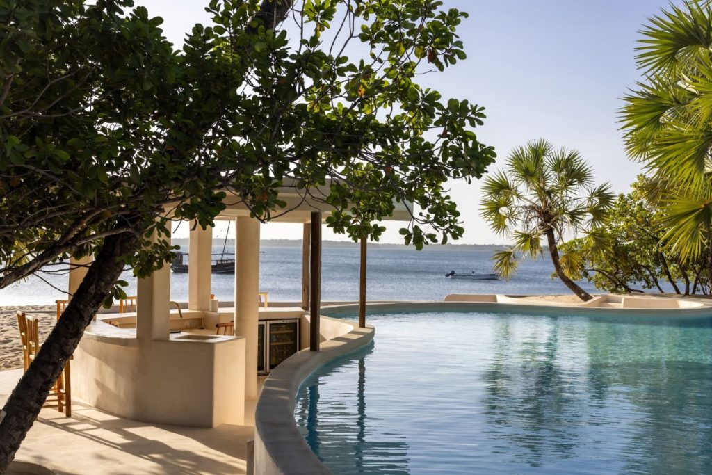 Poolside bar with wooden stools beside a curved pool at The Cabanas Lamu, with a dhow moored in the channel beyond.