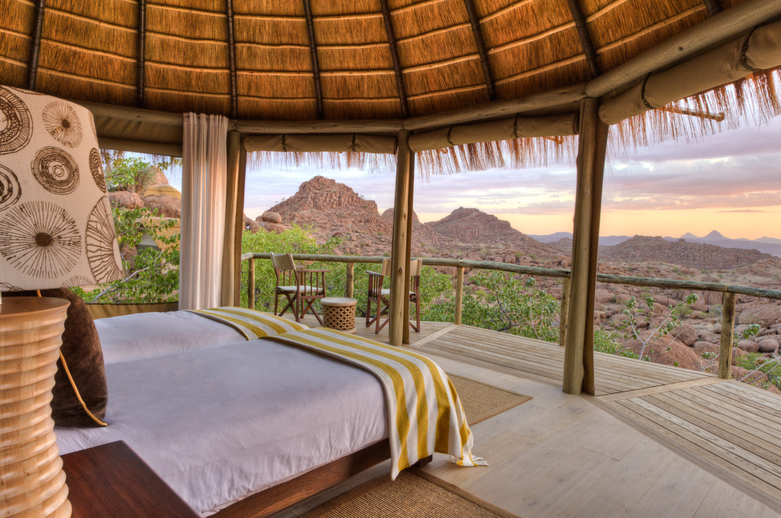 Twin beds beneath a thatched roof open onto a private deck at Mowani, with layered rock formations stretching to the horizon at dusk.