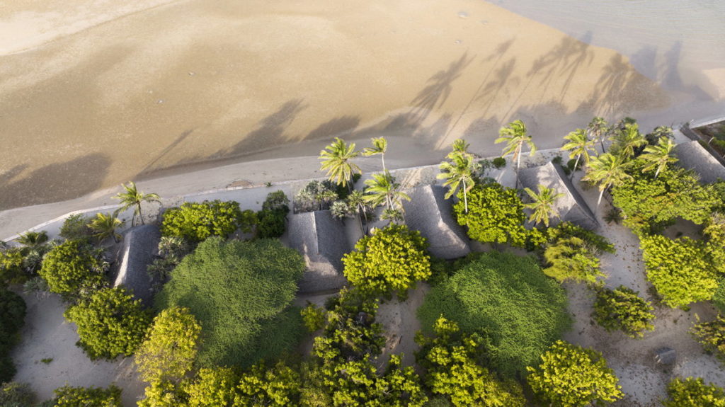 Aerial view of thatched beach cottages among green trees and palms, their long shadows falling across the sand and shallows at low tide.