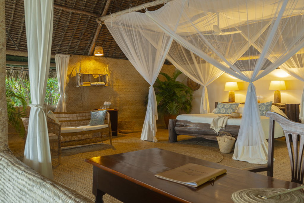 Bedroom at Manda Bay Lodge with a mosquito-netted log bed, woven palm walls, a wicker sofa and warm lamplight under a makuti thatched roof.
