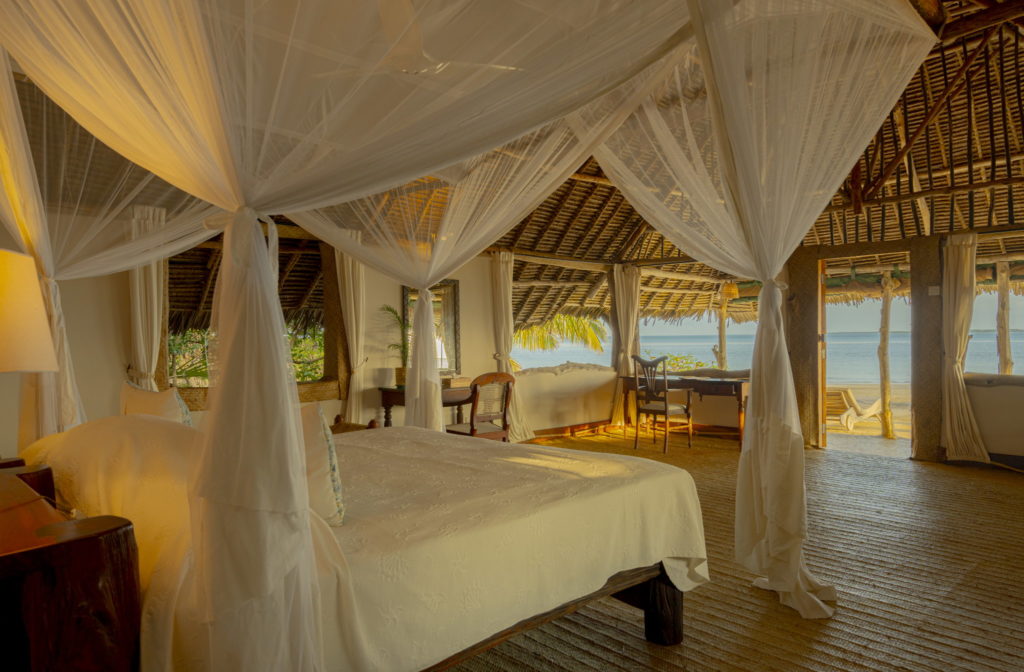 Beachfront bedroom with a canopied bed, writing desk and open doors to the sand and ocean in soft golden light, Lamu Archipelago.