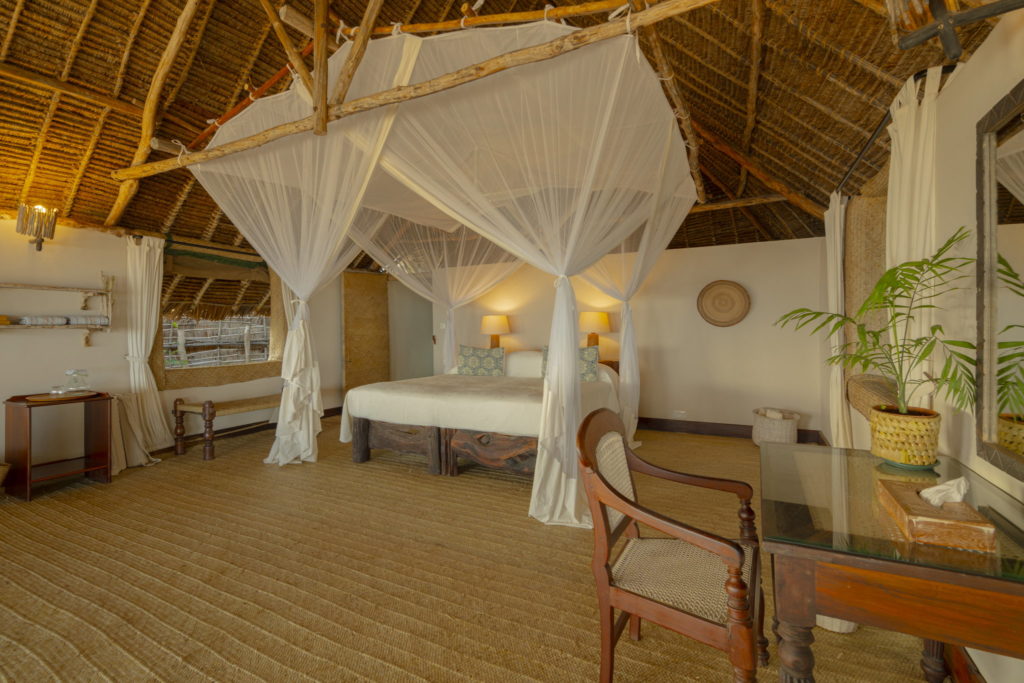 Spacious bedroom with a mosquito-netted double bed on a rustic log frame, coir matting, a wooden writing desk and a high thatched ceiling.