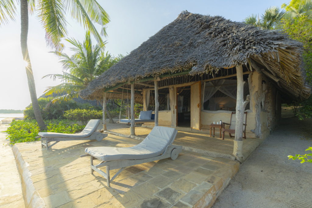 Two cushioned loungers on a stone terrace outside a thatched beach cottage at Manda Bay Lodge, with palms and morning sun behind.