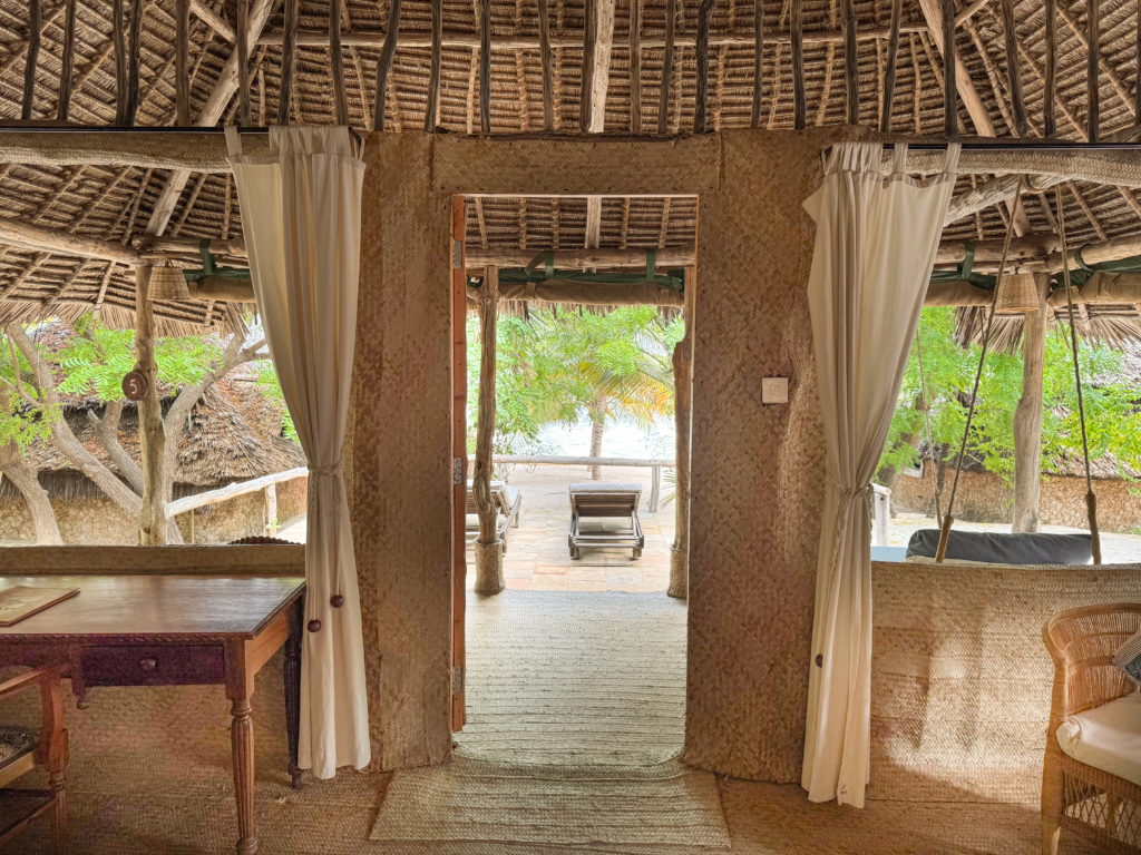 View from a bedroom through an open doorway to a shaded veranda, wooden lounger and palms, between woven palm walls and white curtains.
