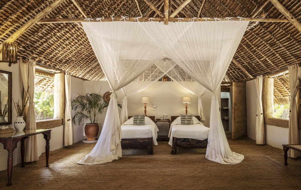Twin room at Manda Bay Lodge with two mosquito-netted beds, bedside lamps, a potted palm and woven palm walls beneath a thatched roof.