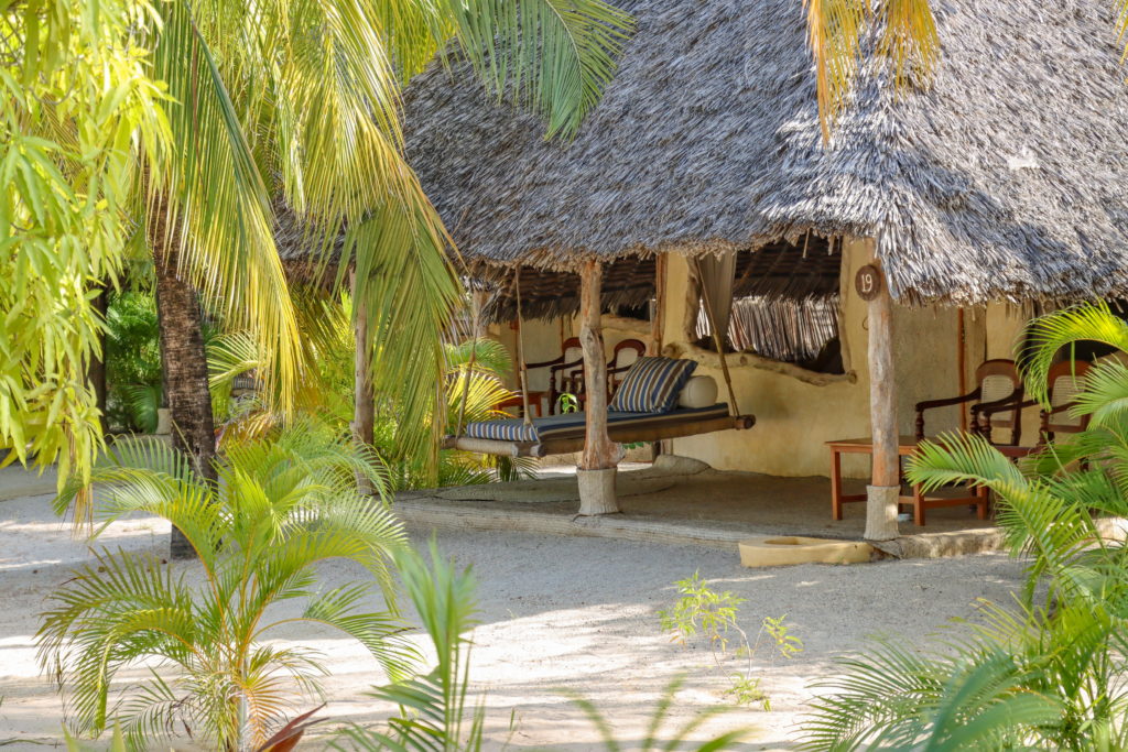 Rope-hung swing daybed with striped cushions on the veranda of a thatched cottage, framed by palm fronds in bright daylight.