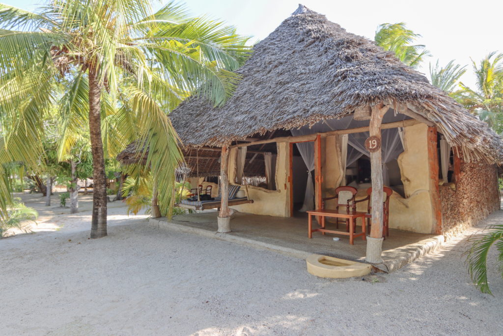 Thatched beach cottage at Manda Bay Lodge with a swing daybed, wooden chairs and a stone footbath on the veranda, shaded by coconut palms.