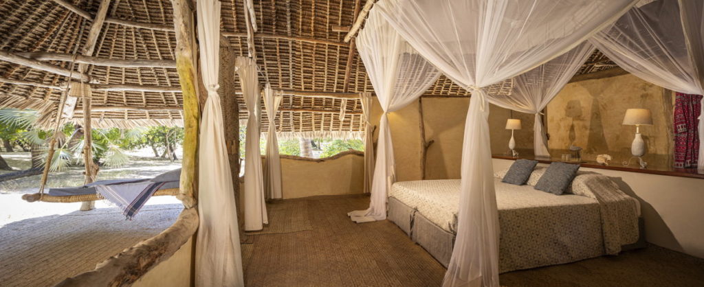 Open-sided bedroom with a canopied bed, white curtains and a swing daybed looking out over sand and palms, Manda Island, Kenya.