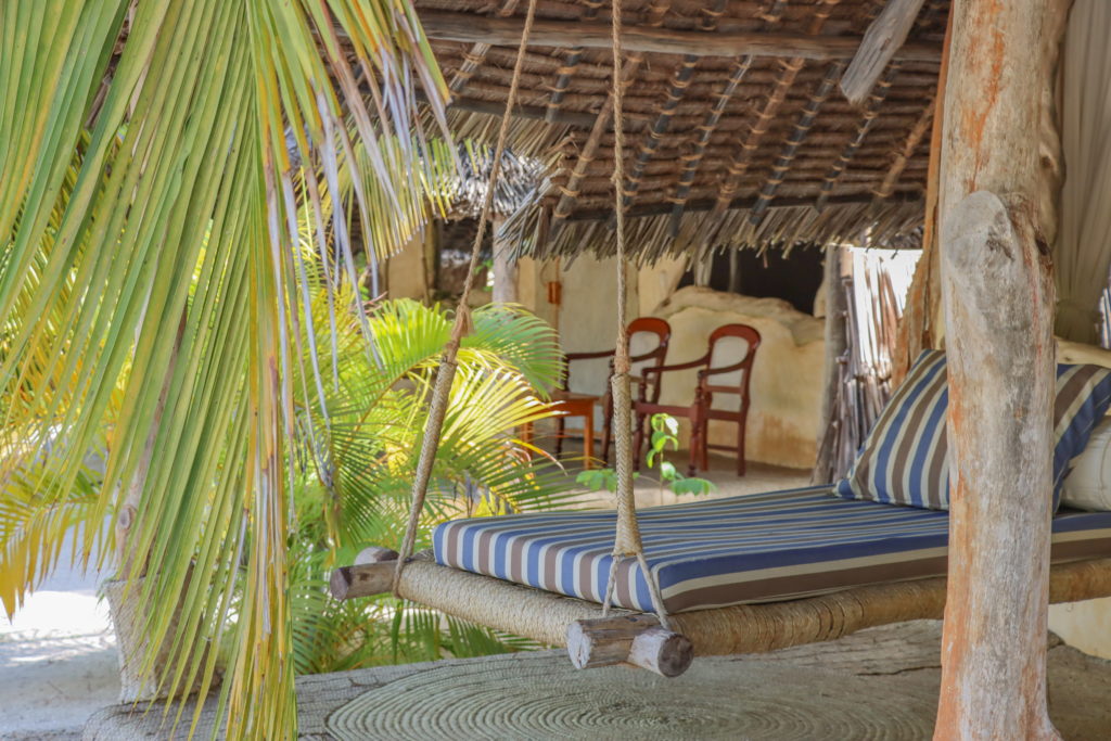 Close-up of a rope swing daybed with striped cushions under a thatched veranda, with wooden armchairs and palm fronds behind.
