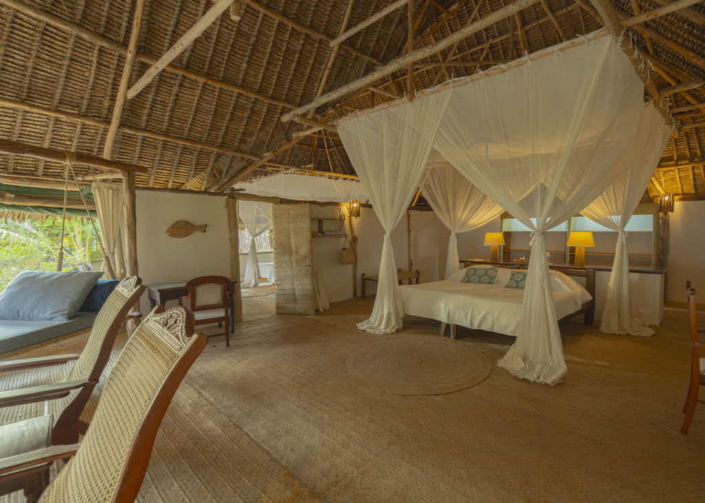 Large bedroom with a canopied double bed, round jute rug, carved armchairs and a cushioned swing daybed under a high thatched roof.