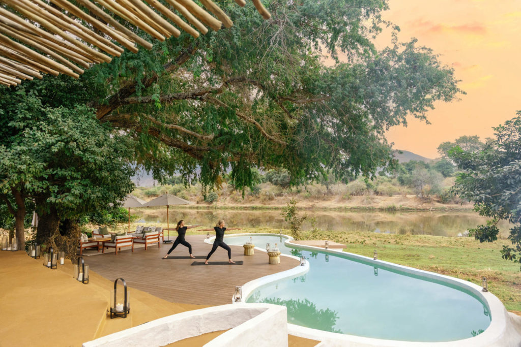 Two guests practise yoga on a deck beside the pool, overlooking the Chongwe River at sunset.