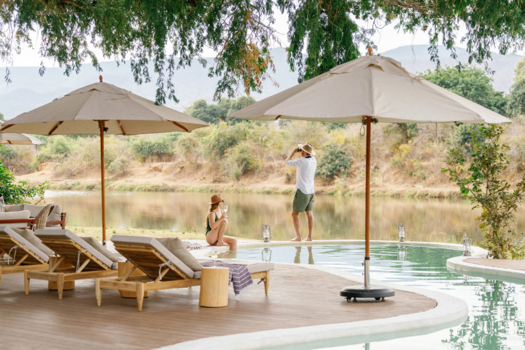 A couple relax by the curved swimming pool, gazing out across the river towards distant mountains.