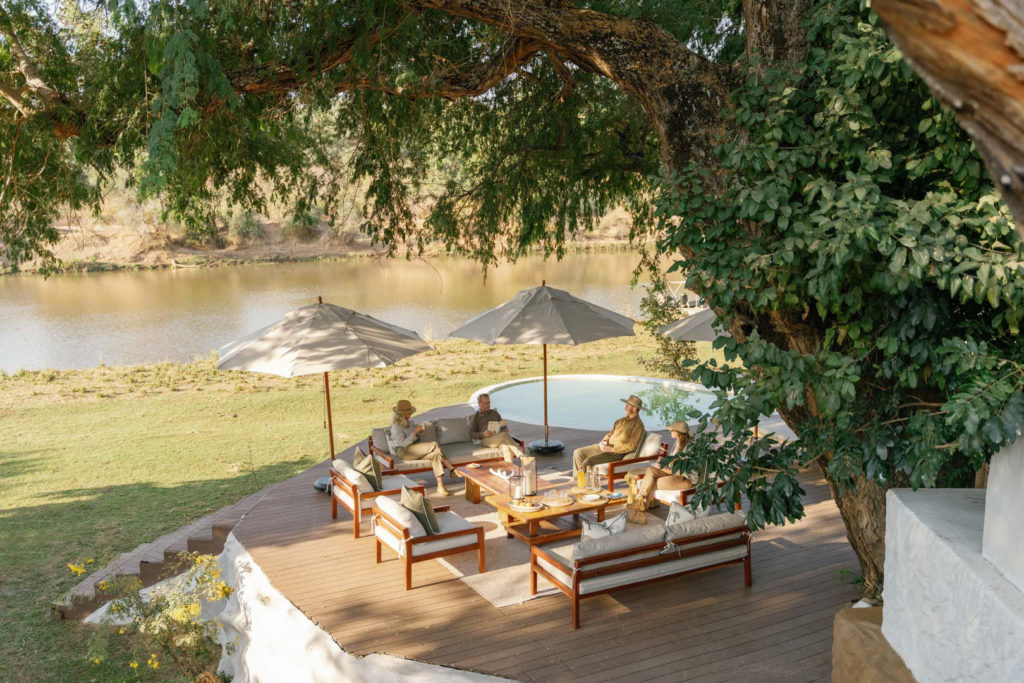Guests gather for afternoon refreshments on a shaded riverside deck at Time + Tide Chongwe House.