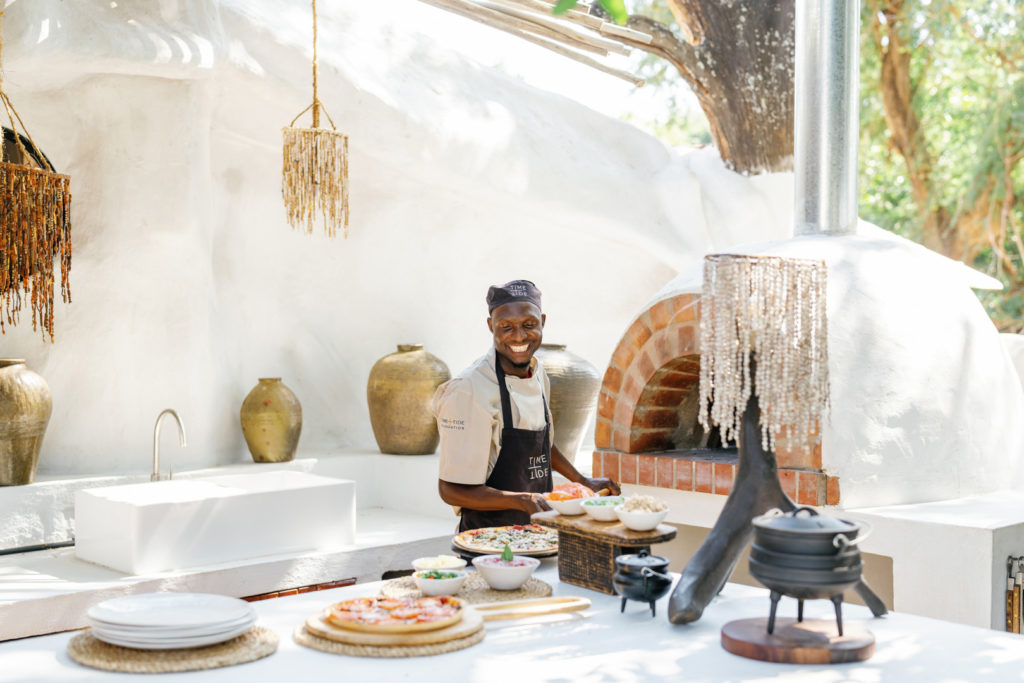 A chef prepares pizzas at the outdoor kitchen's brick oven, framed by hand-crafted beaded chandeliers.