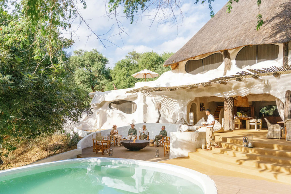 Guests unwind around the boma fire-pit beside the pool, with the thatched house rising behind them.